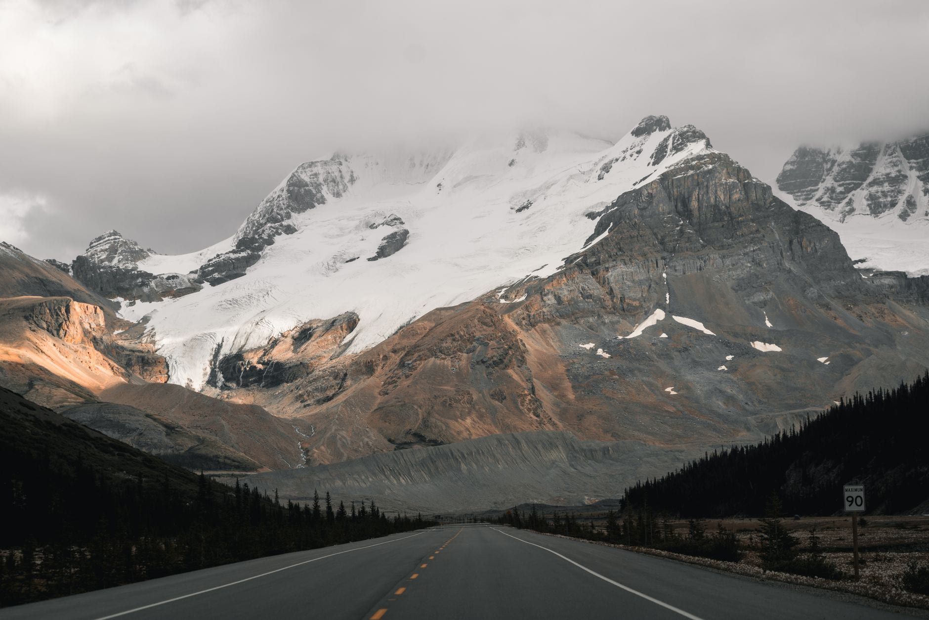 A paved highway stretches toward a towering snow-dusted mountain and hanging glacier under cloudy skies in Jasper National Park.