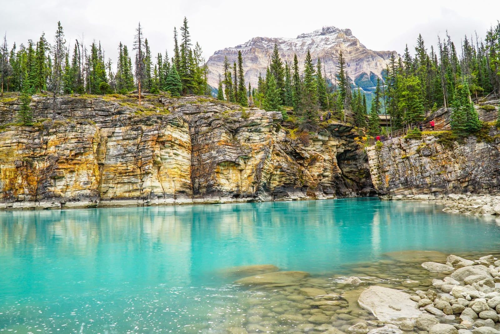 Turquoise water rests below layered rocky canyon cliffs and evergreen trees with a mountain peak in Jasper National Park.