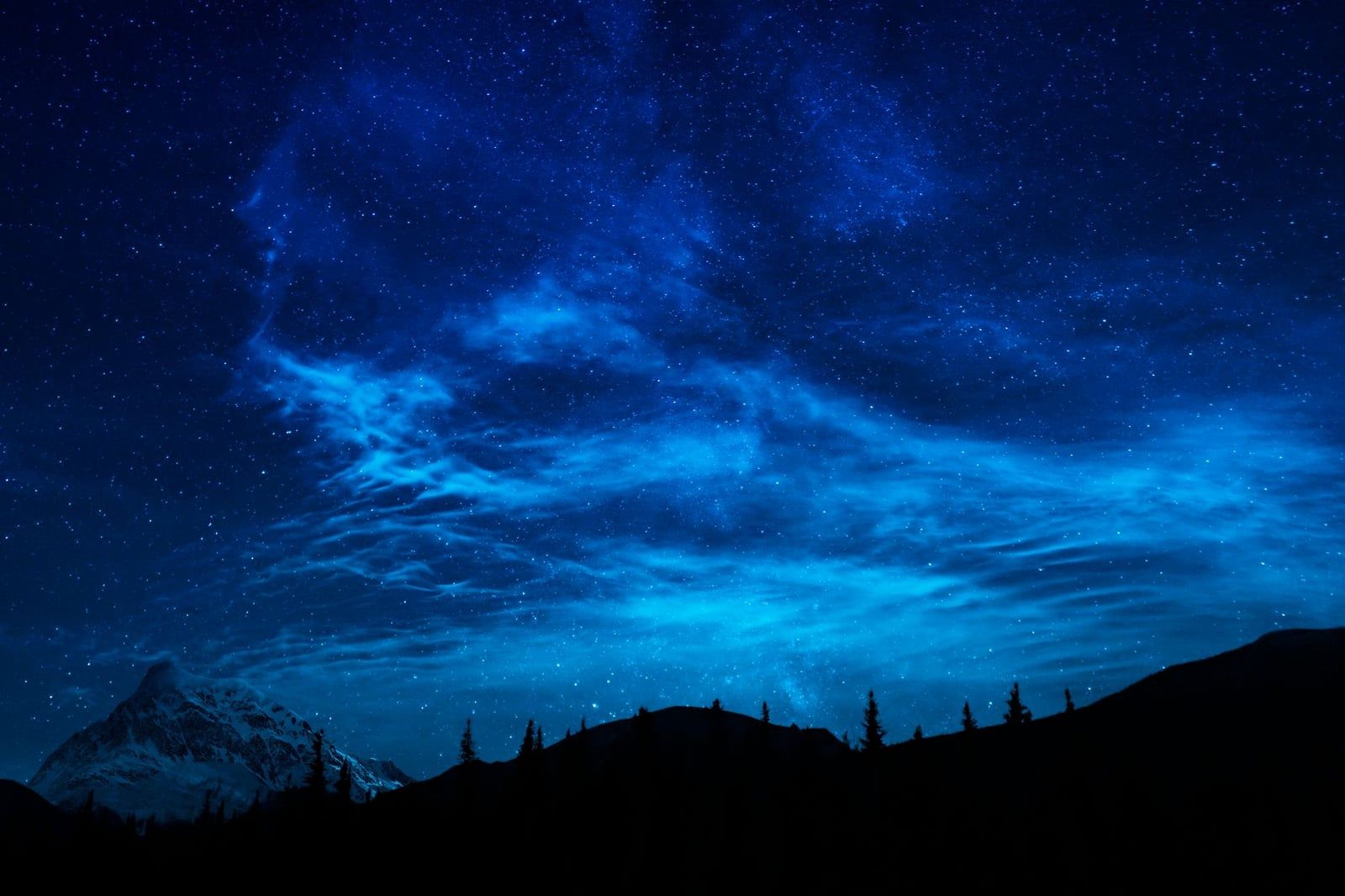 A star-filled night sky and glowing blue clouds over mountain silhouettes in Jasper National Park.
