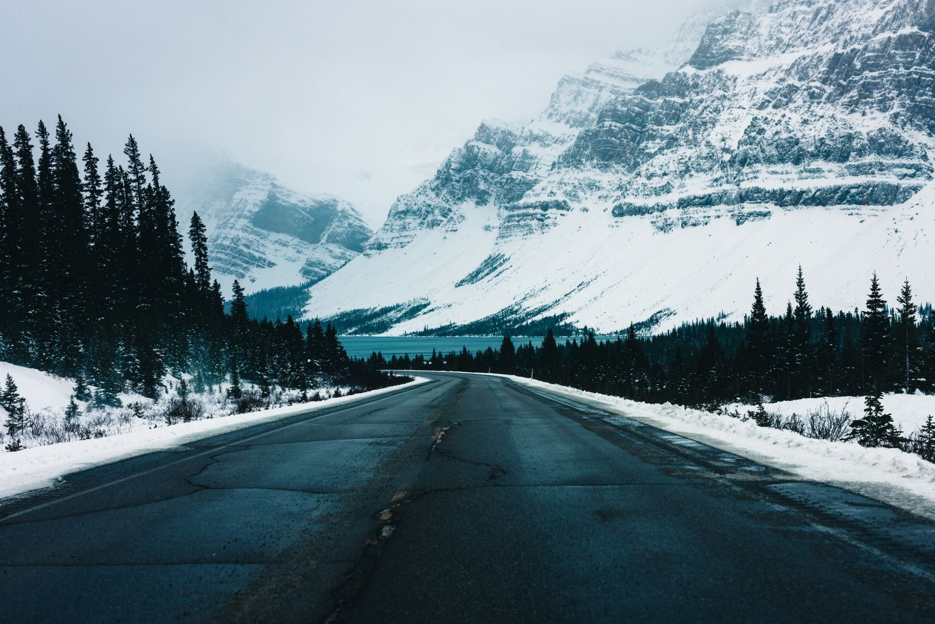 A paved highway cuts through snowy evergreen forests toward a lake and snow-covered mountains in Jasper National Park.