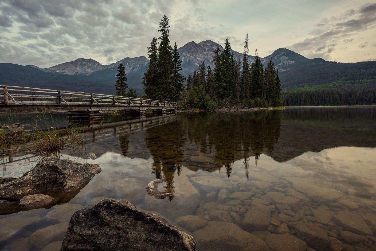 A wooden footbridge leads across Pyramid Lake to forested Pyramid Island, with Pyramid Mountain reflected in the calm water in Jasper National Park.