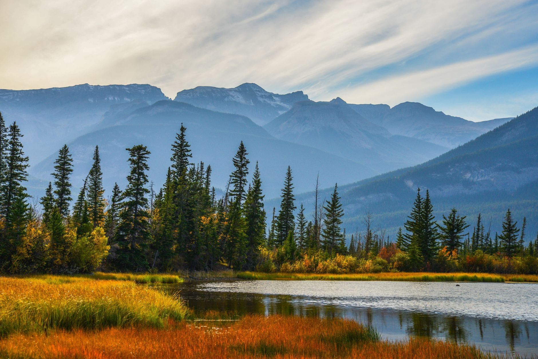 Autumn foliage and pine trees border a quiet lake against a backdrop of mountains in Jasper National Park.