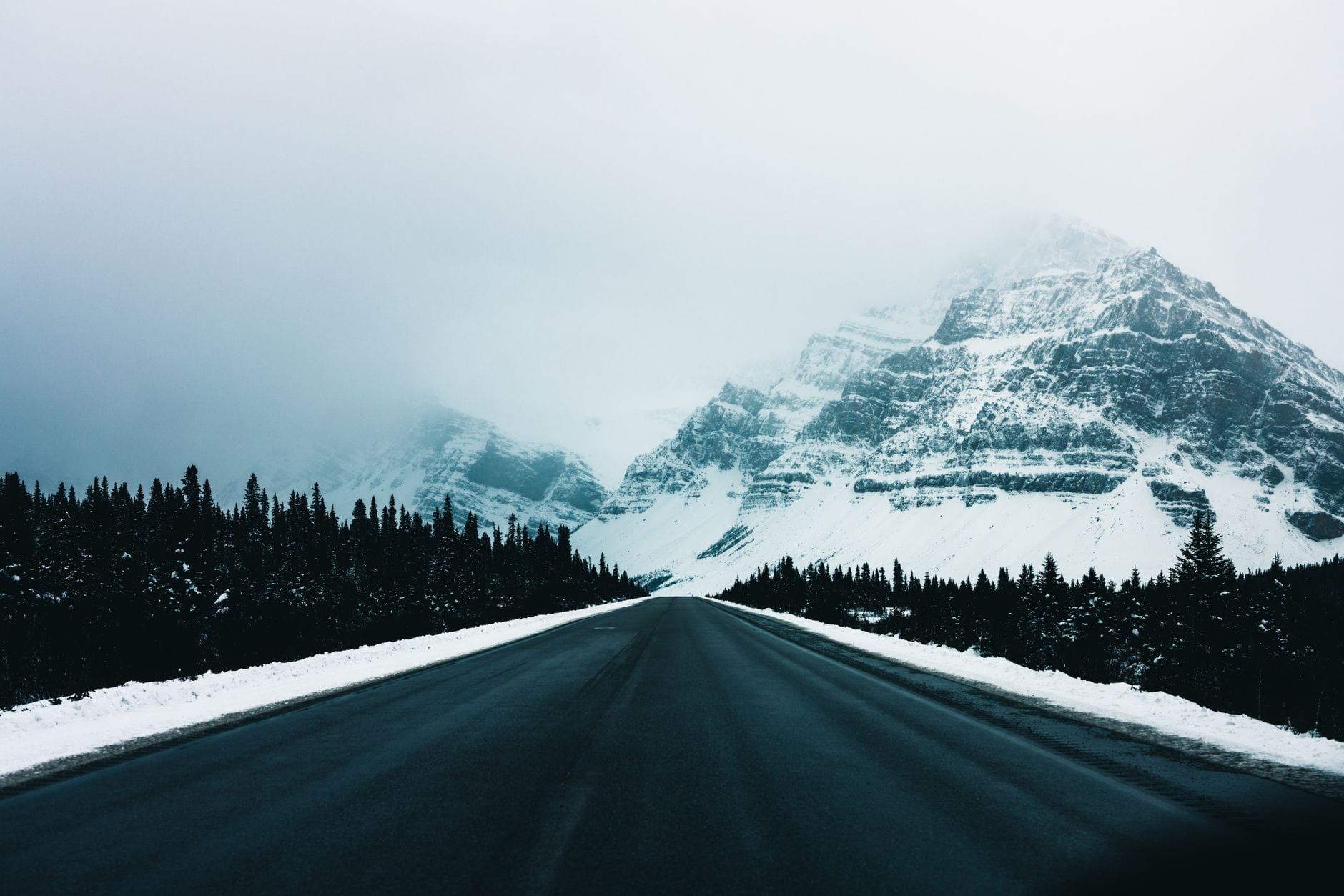 A wide paved highway lined with evergreen trees leads toward mist-shrouded, snow-covered mountains in Jasper National Park.