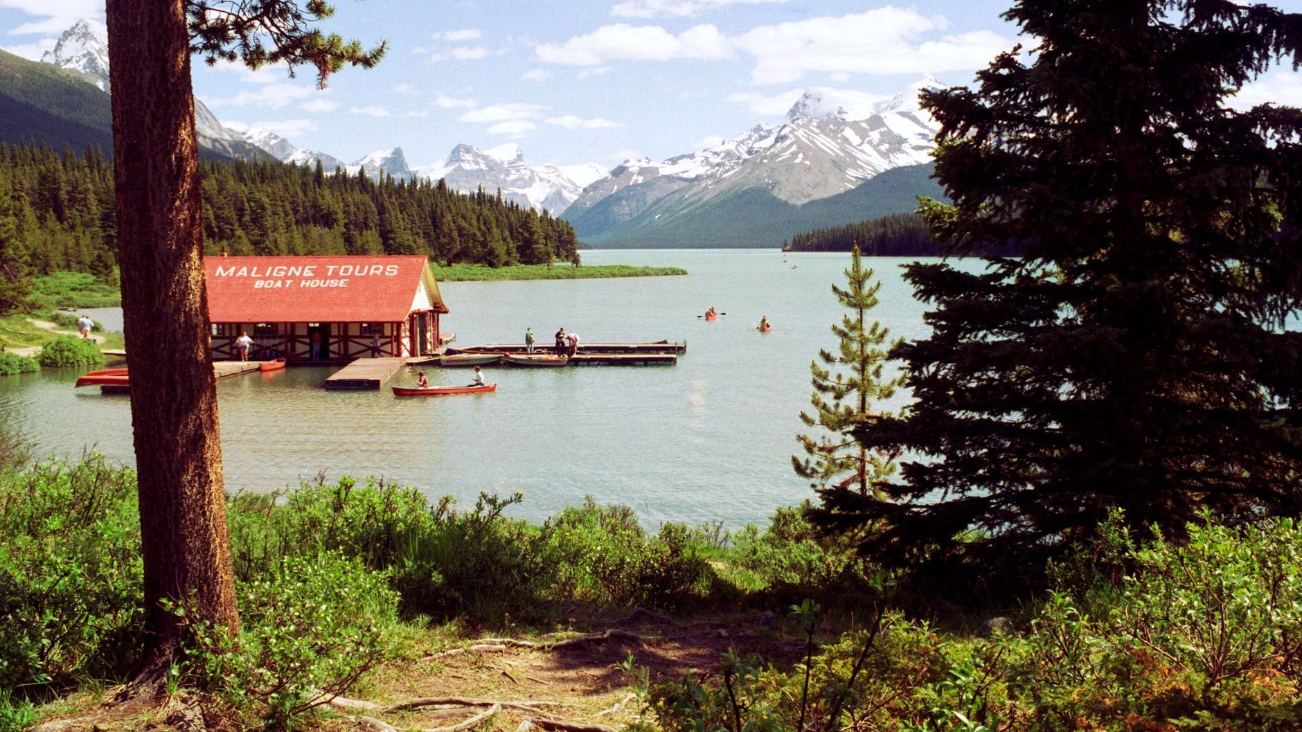 Historisches Bootshaus mit rotem Dach und Kanus auf dem Maligne Lake, umgeben von Nadelwäldern und schneebedeckten Gipfeln im Jasper-Nationalpark.